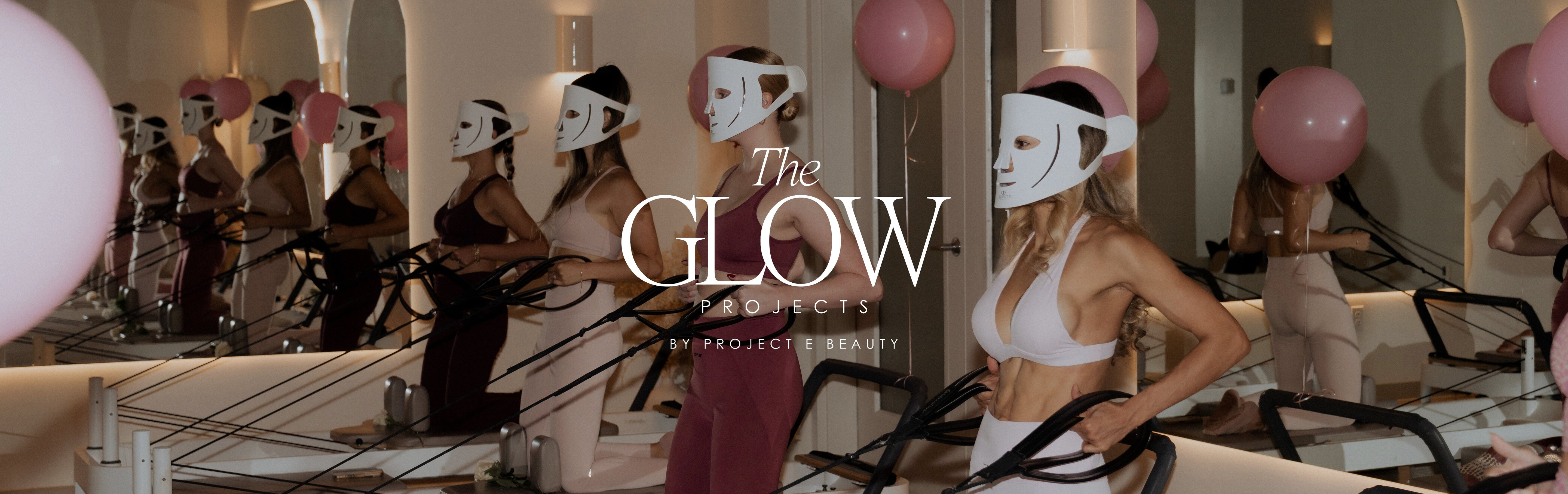 Women in Pilates class wearing LED red light therapy masks at The Glow Projects by Project E Beauty, stylish activewear in white, pink, and burgundy, with pink balloons in mirrored studio.