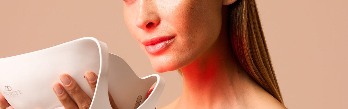 Woman holding the LumaLux Face V2 LED mask beside her face with red light glowing